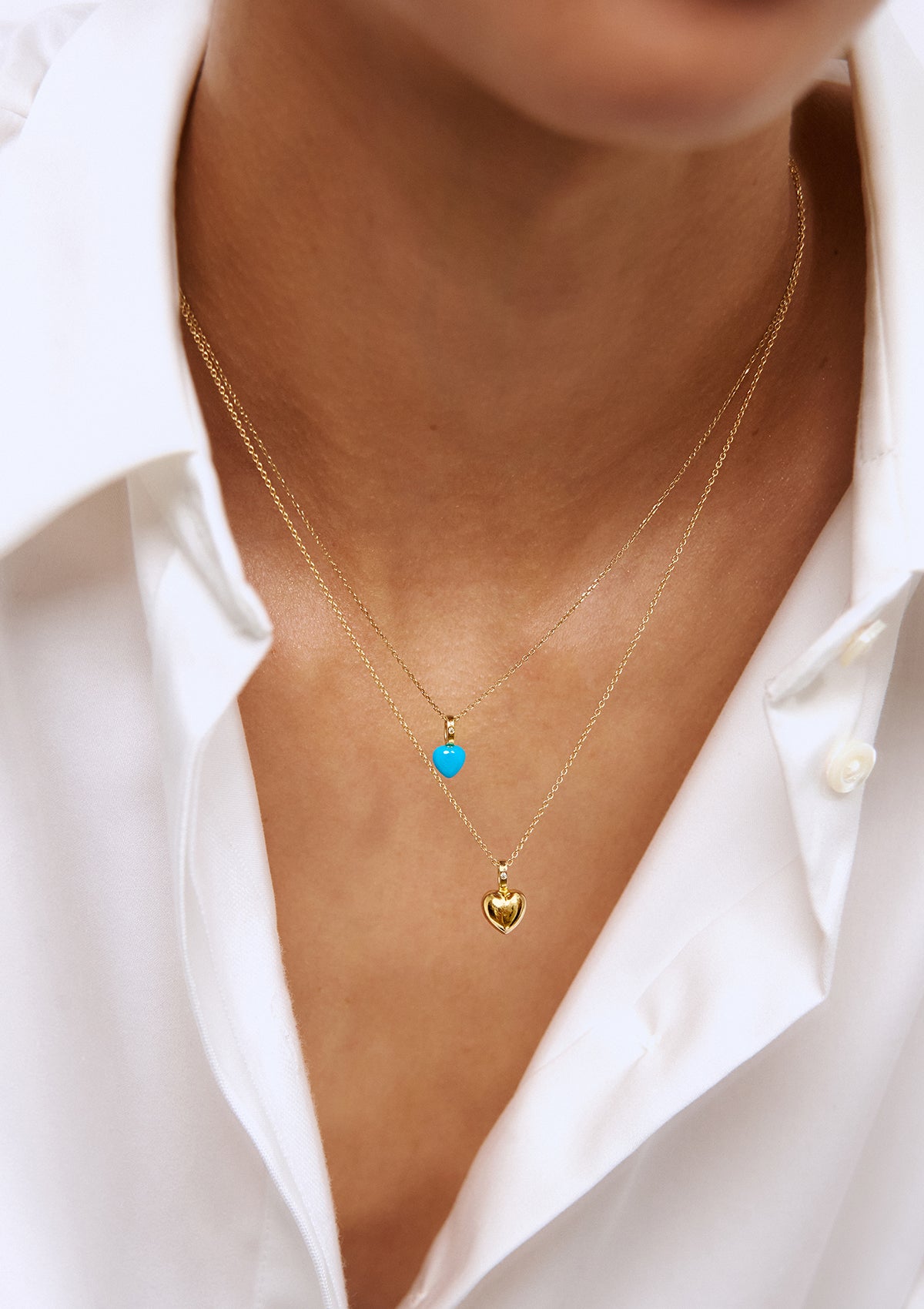 woman in a white dress shirt wearing two of our Heart pendants. One of the pendants is a heart shaped turqouise gemstone and a diamond, and the second one is made in solid gold set with a diamond.