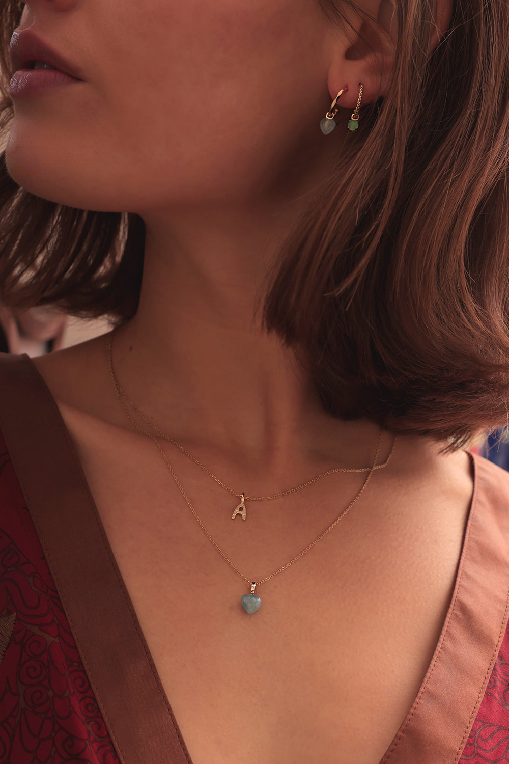 Woman with a gold pendant set with a heart-shaped Aquamarine gemstone and a diamond. Also wearing a letter pendant in solid gold set with Diamonds with the letter 'A'