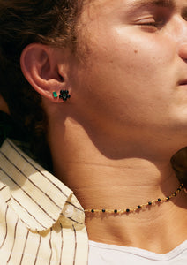 A young man laying in the sun wearing flower shaped earrings with black and green gemstones and a black agate necklace